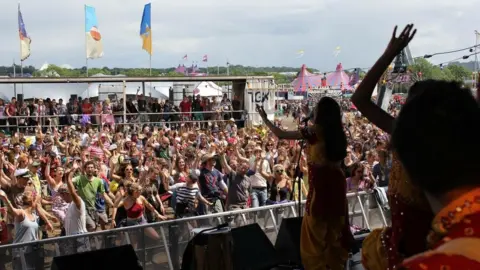 Eternal Taal Women wearing yellow and red sequinned outfits are on a stage in front of a large grown of people with their hands in the air. They are dancing and singing 