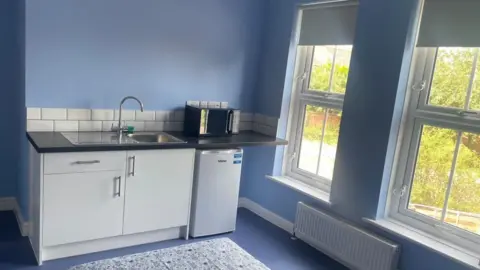 Pictured is a room. On the far wall is a kitchen sink with two cupboards and a drawer underneath. On the right is a microwave on a counter top and under that is a white fridge. The walls are painted bright blue and through two windows, you can see trees.
