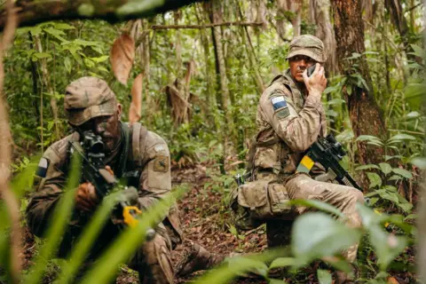 British Army Two army soldiers armed with machine guns and dressed in camouflage crouching in the jungle while one tests a new smartphone.