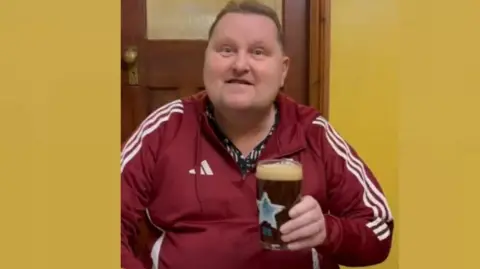 John Booth John Booth, wearing red Adidas tracksuit, and holding pint glass. He is looking at the camera and has short, light brown hair. The glass has the blue star logo of Newcastle Brown Ale.