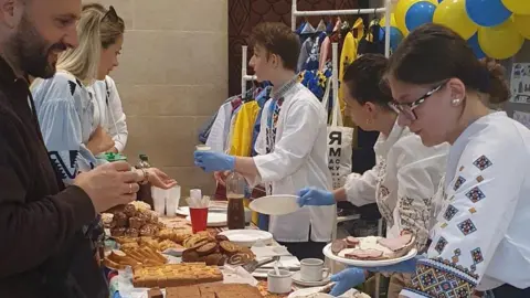 Antonina Grebeniuk People wearing white outfits serving food to others with yellow and blue balloons behind them