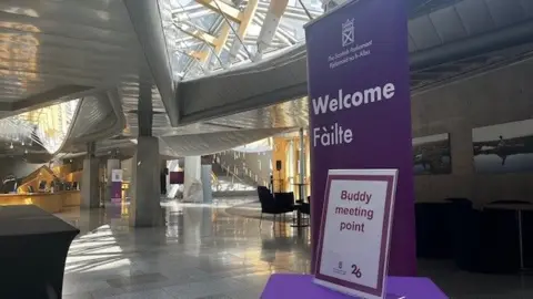 The lobby of the Scottish Parliament, a light grey building with lots of wood. There is a purple sign welcoming people in Gaelic and English and a sign saying "Buddy Meeting Point".