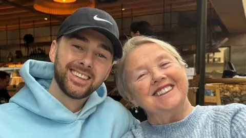 (L-R) A son and mum. The son is wearing a blue hoodie and a black sun hat. The mum is wearing a grey/blue sweater. They are both smiling at the camera and appear to be sitting in a cafe. 