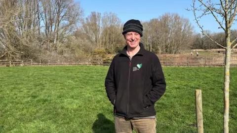 Adrian Harms / BBC A man in a black fleece stood in a field. He has his hands in his pockets and is smiling.