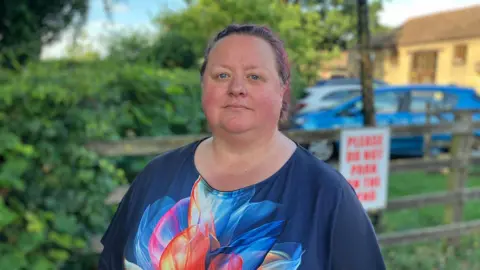 Sam Read/BBC Charlotte looks at the camera while standing outdoors near a wooden fence. There are bushes and a building behind, and she wears a blue T-shirt with an orange and blue pattern on. Her hair is pinky brown and tied back.