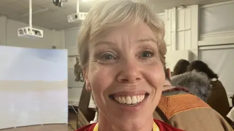 Lara King Woman with blonde hair wearing a red top smiling. There are people behind her and a screen