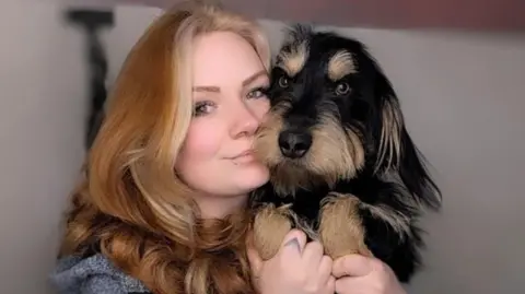 Jody Hall Jody Hall cuddling her dog, Chester, who is black and tan with long fur and fluffy brown eyebrows. Jody has long blonde and auburn hair.