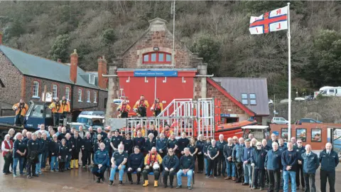 A photograph of a large group of people posing outside a brick building and a flag pole, looking at the camera. Almost all of them are in RNLI uniforms. There is an RNLI flag on the pole, and the building is backed by trees.