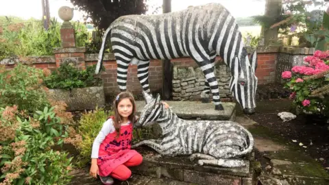 Clare Stephens A little girl with long brown hair in a pink top and trousers crouches down next to a statue of a zebra with another larger zebra statue behind her