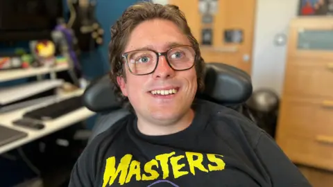 A person sits in a wheelchair, wearing a black T-shirt with the word 'MASTERS' in yellow. Behind them, a room contains a guitar mounted on the wall, shelves, and a desk with computer equipment.