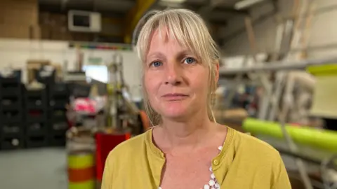 Laura Jones has blonde hair tied back and is wearing a yellow top with buttons. A white crafted shell necklace is just visible.