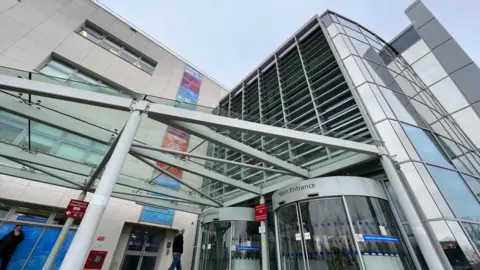 The main entrance of Broomfield Hospital in Chelmsford showing a metal and glass canopy sticking out from the front of the building which is clad with a natural coloured material with multi-coloured blocks forming a strip down the middle of the building. There are rotating doors.