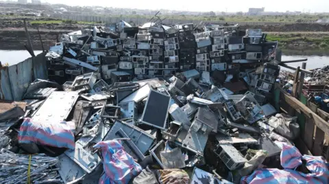 Tumpukan besar limbah elektronik, termasuk layar besar, di tempat pembuangan sampah Agbobloshie. Perairan terlihat di latar belakang