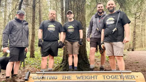 Five men are stood in a line behind in the woods. At their feet is a log with the words carrying the weight together printed on