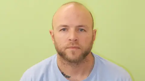 Kirk Bishop - A man with a bald head and a stubbly brown beard and moustache sitting with a grey t-shirt on. He is sitting in front of a green wall. On his neck you can see the wording from a tattoo. 