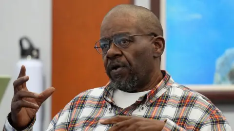 A man wearing a checkered shirt and glasses testifies in court.