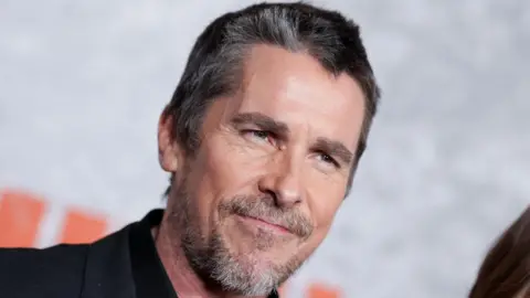 Headshot of Bale. He has greying black hair. He looks past the camera