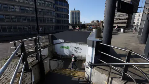 A man walks down some concrete steps into a subway entrance. There is graffiti on a wall behind him. At ground level, a number of buildings can be seen.