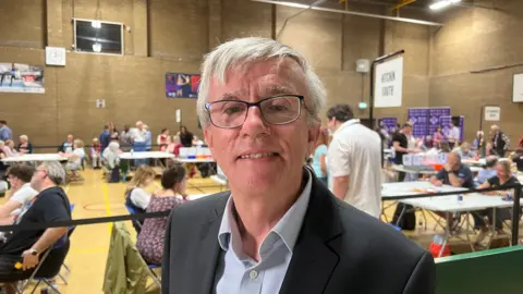 Simon Dedman/BBC A person wearing glasses, a light-coloured shirt and a dark jacket stands in the foreground of a busy vote-counting venue. Behind them, election workers and observers sit and stand around tables covered with ballot papers, while constituency signs hang from the sports hall walls.