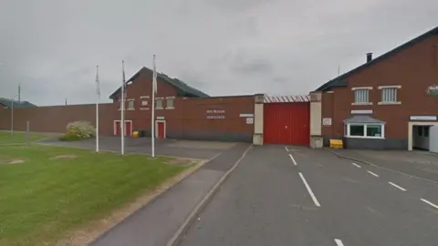 The entrance to Dovegate prison with a height restricted red gate topped with wire between two buildings. There is an area of grass and flags on the left side of the gate.