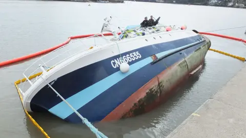 UK government A French trawler capsized in Dartmouth Harbour. The boat is blue and white. An orange inflatable boom surrounds the vessel. Two people are stood on part of the boat.