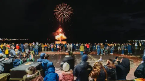 Hundreds of people in warm coats and hats stand on Douglas promenade looking out at the bay, which has red fireworks exploding.
