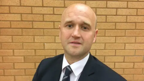 A picture of Dafydd Llywelyn stood in front of a brick wall. He is wearing a suit and tie.