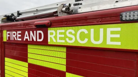 BBC The side of a fire engine with red and green/yellow rectangles in a pattern and "fire and rescue" written on the side.
