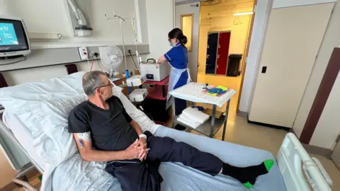 Tommy Richards preparing for the re-infusion of his CAR-T cells in a ward at Addenbrooke's Hospital. He is sitting upright in a bed looking towards a staff member, who has her hands inside a piece of machinery. He has short grey hair and glasses, and is wearing a grey T-shirt, navy jogging bottoms and black and green socks. The worker has brown hair in a pony tail, and is wearing a face mask and a white disposable apron over royal blue scrubs.