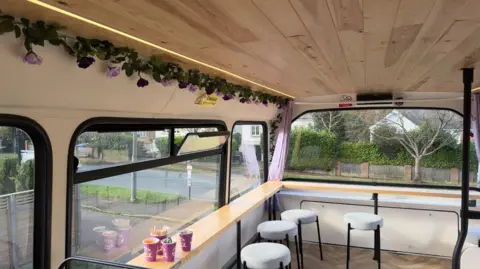 Top deck of double decker, decorated with flowers with stools and tables and a view out of the front window.