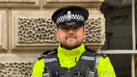 George King/BBC A close-up head and shoulders image of PC Lewis Finch. He has a dark beard and is smiling. He is wearing a police uniform and looking straight into the camera.
