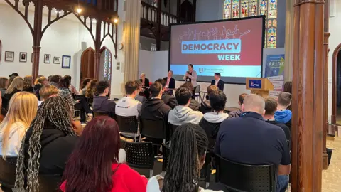 Four people are on a stage in front of a projector which says Demoracy Week. One woman is standing and speaking into a microphone while the others are seated. Students are sat on black chairs in a hall looking at the politicans. The Hall is a converted church and there's a stained glass window in the background.