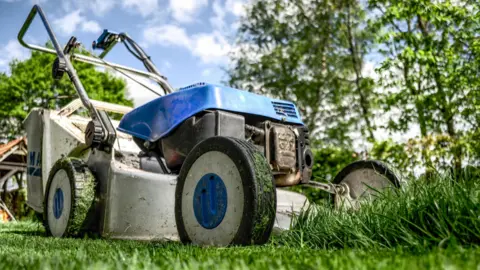 Durham University A blue and grey lawnmower is currently cutting down some long green grass. In the background, tall green trees are visible.