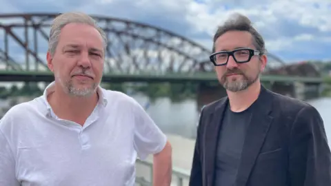 Two men face the camera with neutral expressions. One wears a white polo shirt and the other wears heavy-framed black glasses and a black t-shirt and blazer. The Vysehrad railway bridge's arches are in the background.