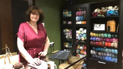 Sharon Cane is pictured standing in her knitting shop. She is standing at the counter with a laptop in front of her. She is facing and smiling at the camera. She has shoulder length brown hair and is wearing a dark red dress. To her right are black coloured shelves holding tens of yarn balls in all different colours.