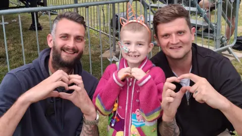 Glen Spooner on the left is wearing a grey hoodie and is looking directly at the camera and smiling. Next to him is Kayla, a girl wearing a pink colourful cardigan. On the right is Owen Spooner, a man wearing a white T-shirt and black zip up jacket. They are all looking directly at the camera and smiling.