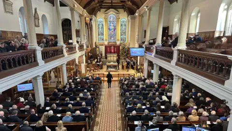 The congregation gathered for the 2024 service from above. There are pillars on either side of the church and large stained glass windows above the alter.