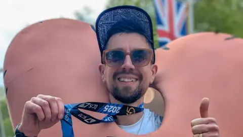 Toby Freeman holding a medal wrapped around his neck and giving a thumbs up to the camera. He is wearing sunglasses and a blue cap and is wearing an outfit designed to look like a pair of large testicles.