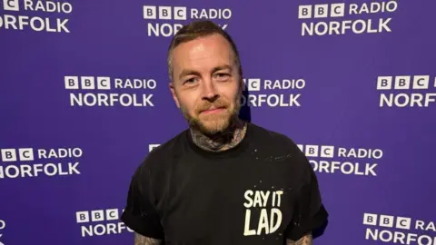 Rob Butler/BBC Jonny Human has short dark hair combed up on top and cropped short on the sides. He has a light brown full beard and moustache. He is wearing a Say It Lad branded tee shirt, and is standing in front of a BBC Radio Norfolk branded wall.