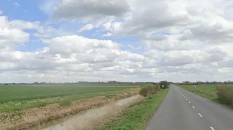 Putney Hill Road, near Prickwillow. It shows a single-carriageway road with a wide water-filled ditch on the left and fields beyond. On the right are more fields. Straight ahead are two national speed limit signs 