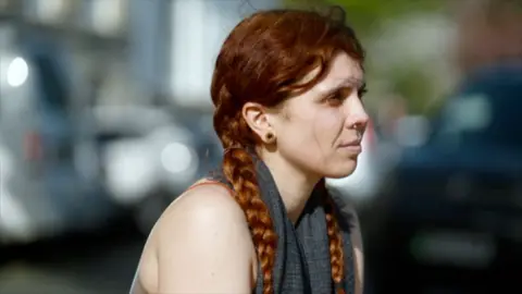 Ellie has red hair in two braids and a nose piercing, she sits outdoors, looking to the side, with blurred traffic in the background.