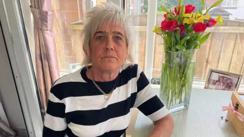A woman with short white hair and a black and white striped top, and a gold necklace, sits at a table with her left arm leaning on it. On the table is a bunch of flowers in a glass vase. A large window is behind her.