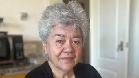 Liz is looking at the camera. She has short grey hair. She is wearing a black cardigan and blue patterned top.