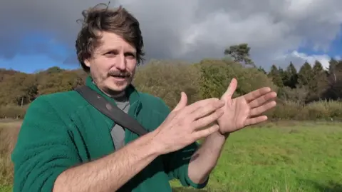 Rob Farrington in a green fleece, standing in an area of green restored wetland, gesturing to his left