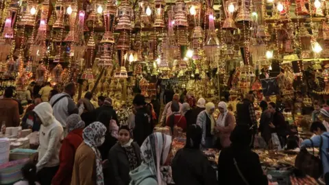 Getty Images Women walk past a market stall with lit up lanterns. The stall area looks quite crowded.