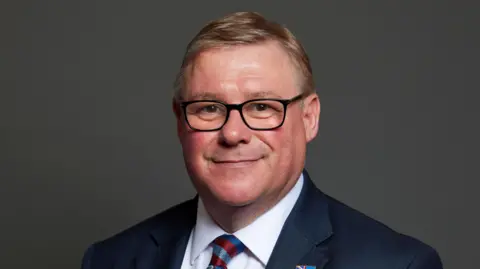 Mark Francois MP's official picture portrait from the House of Commons. He has short brown hair and black glasses and is smiling. He is wearing a blue jacket, a blue shirt and a patterned tie.