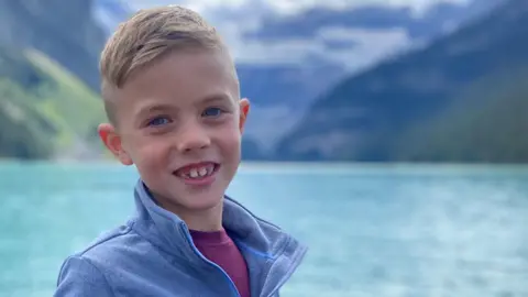 Family handout A young blond boy stands in front of a large body of water and smiles at the camera. He is wearing a dark red t-shirt and a blue sweater over it.