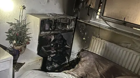 LFB A bedroom damaged by fire. Broken furniture, bedding and walls covered in soot, as well as a small Christmas tree