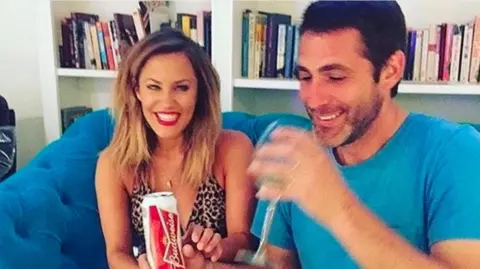 A woman smiles while looking towards a man with a blue shirt taking a drink from a glass while sat on a blue sofa in a room with books on a bookcase behind them
