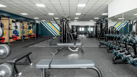 James McCauley A gym with a range of machines in the centre of the room, with a dumbbell rack on the left of the image, and on the right there are three lifting racks with colourful weight plates on the sides of the racks. At the bottom of the picture are two gym benches.